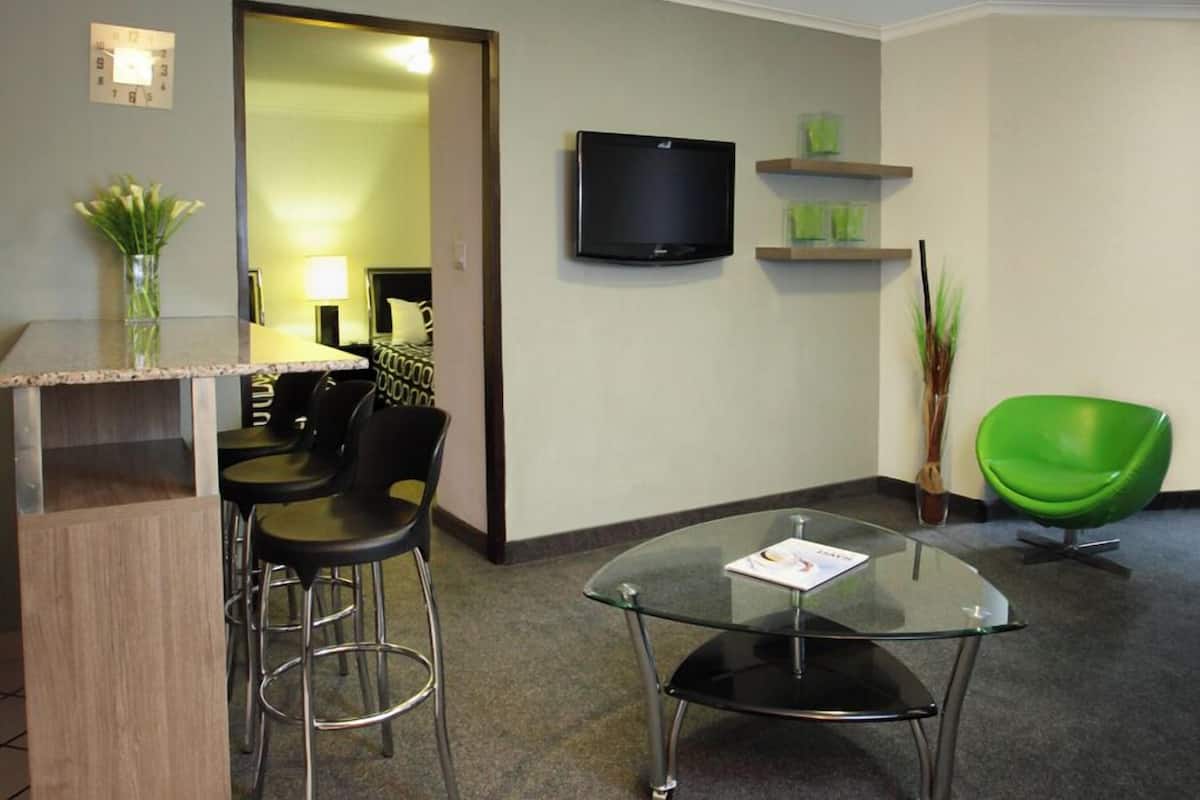 A hotel room with a bar area, a television, a green chair, and a glass table.