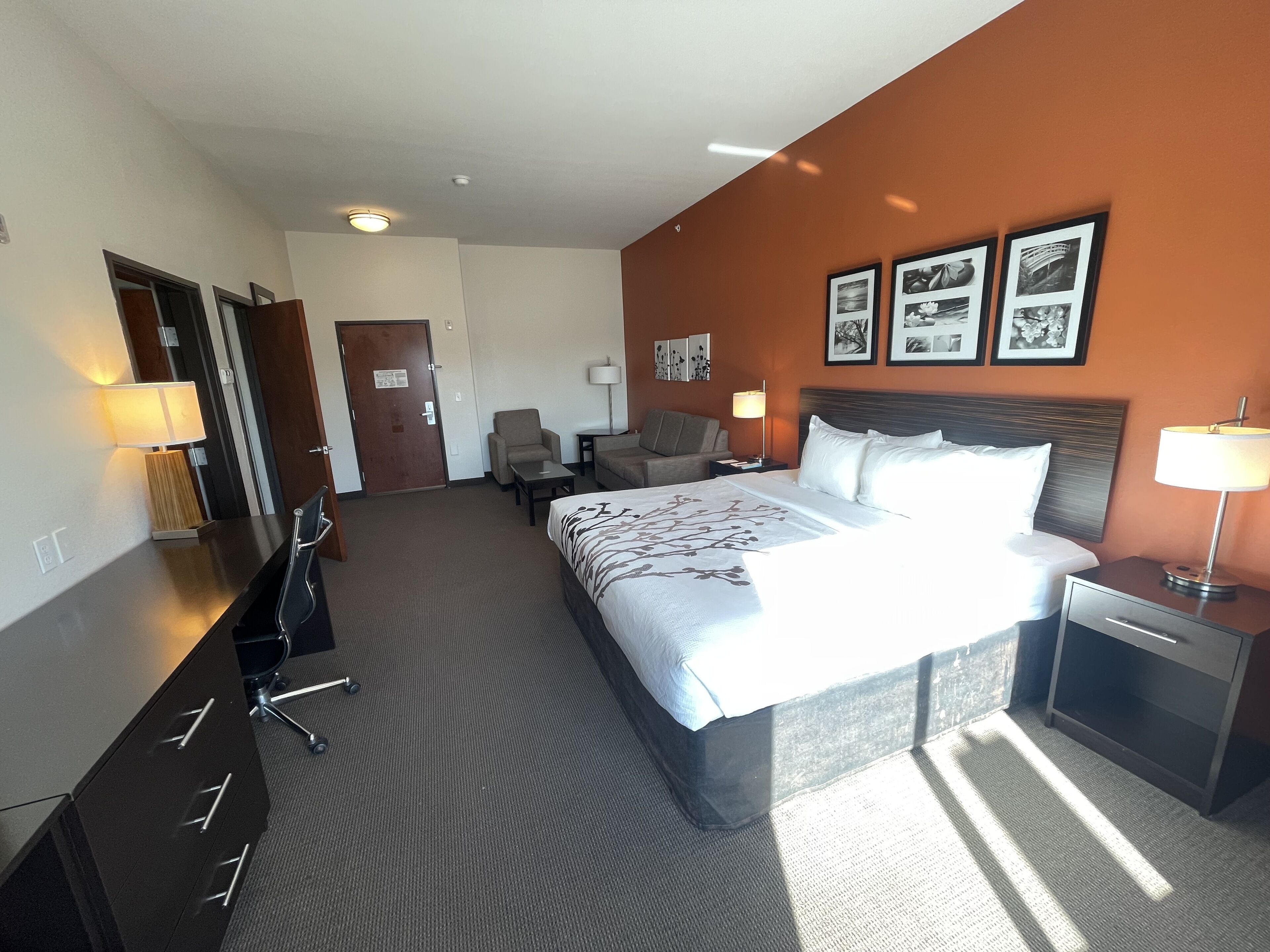 A hotel room with a large bed, a desk, a chair, a small table, and a lamp.