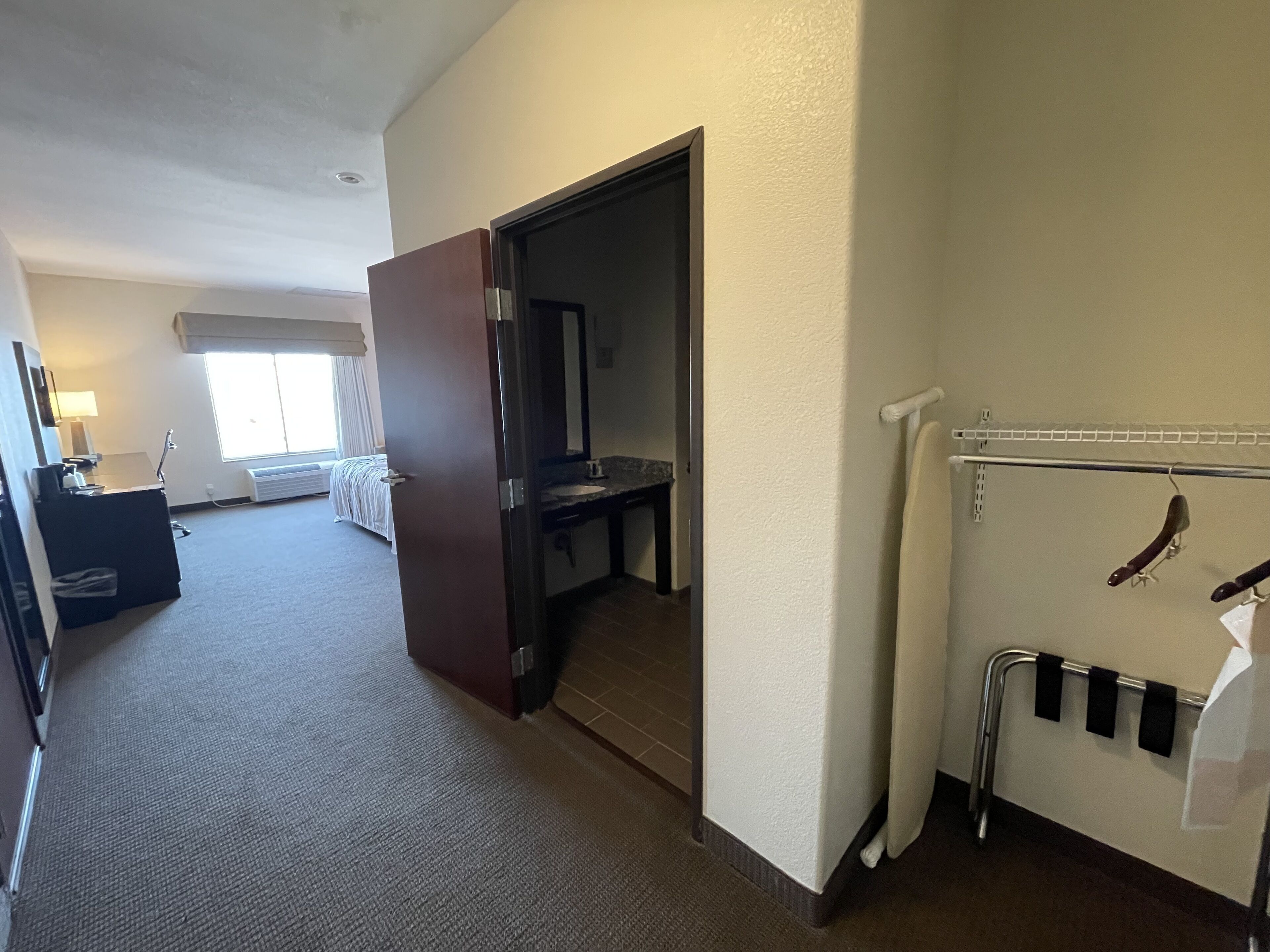 A hotel room with a bed, a desk, a chair, a window, and a bathroom with a shower and towel rack.