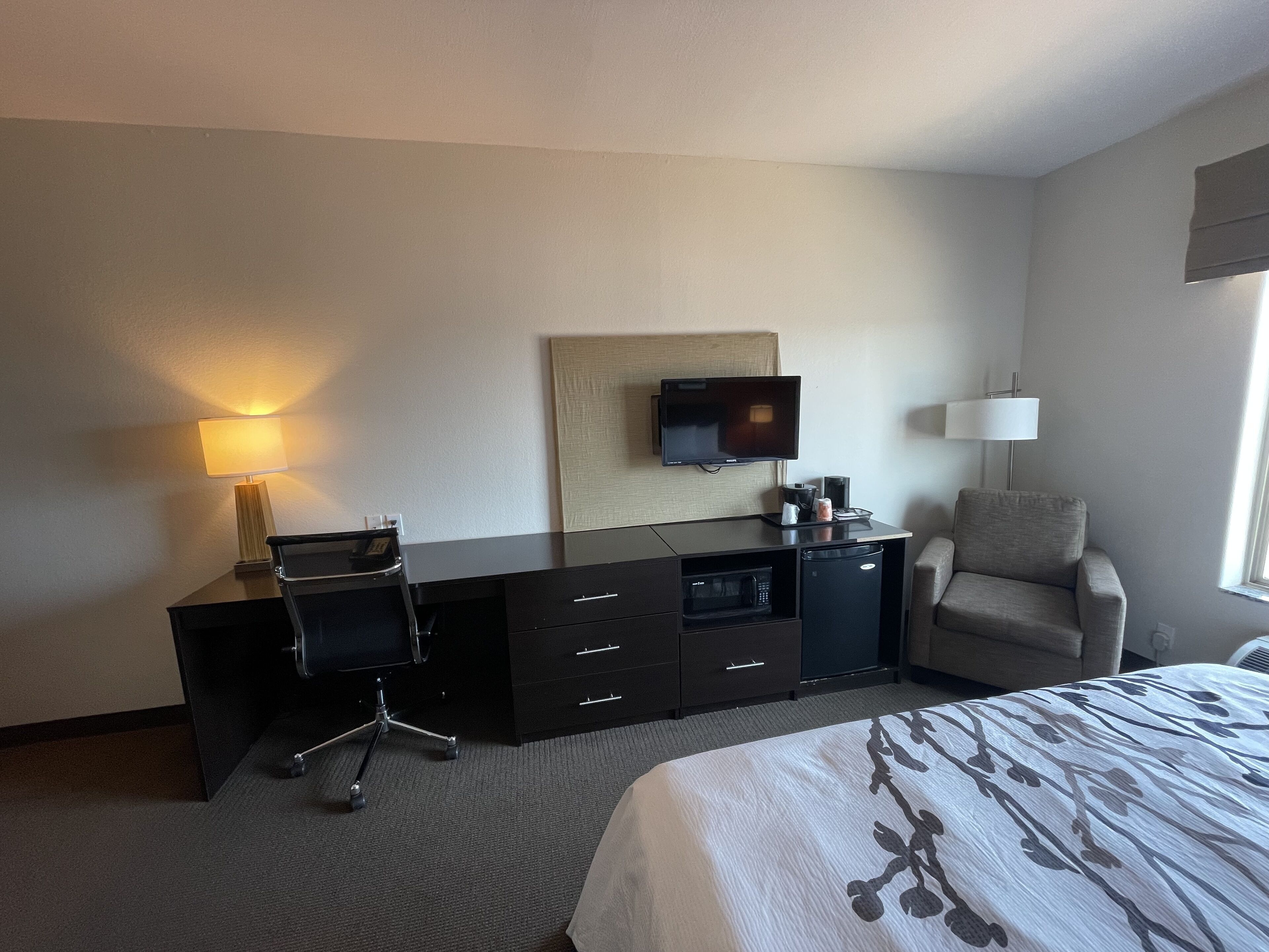 A hotel room with a bed, a desk with a chair, a television, a lamp, and a chair.
