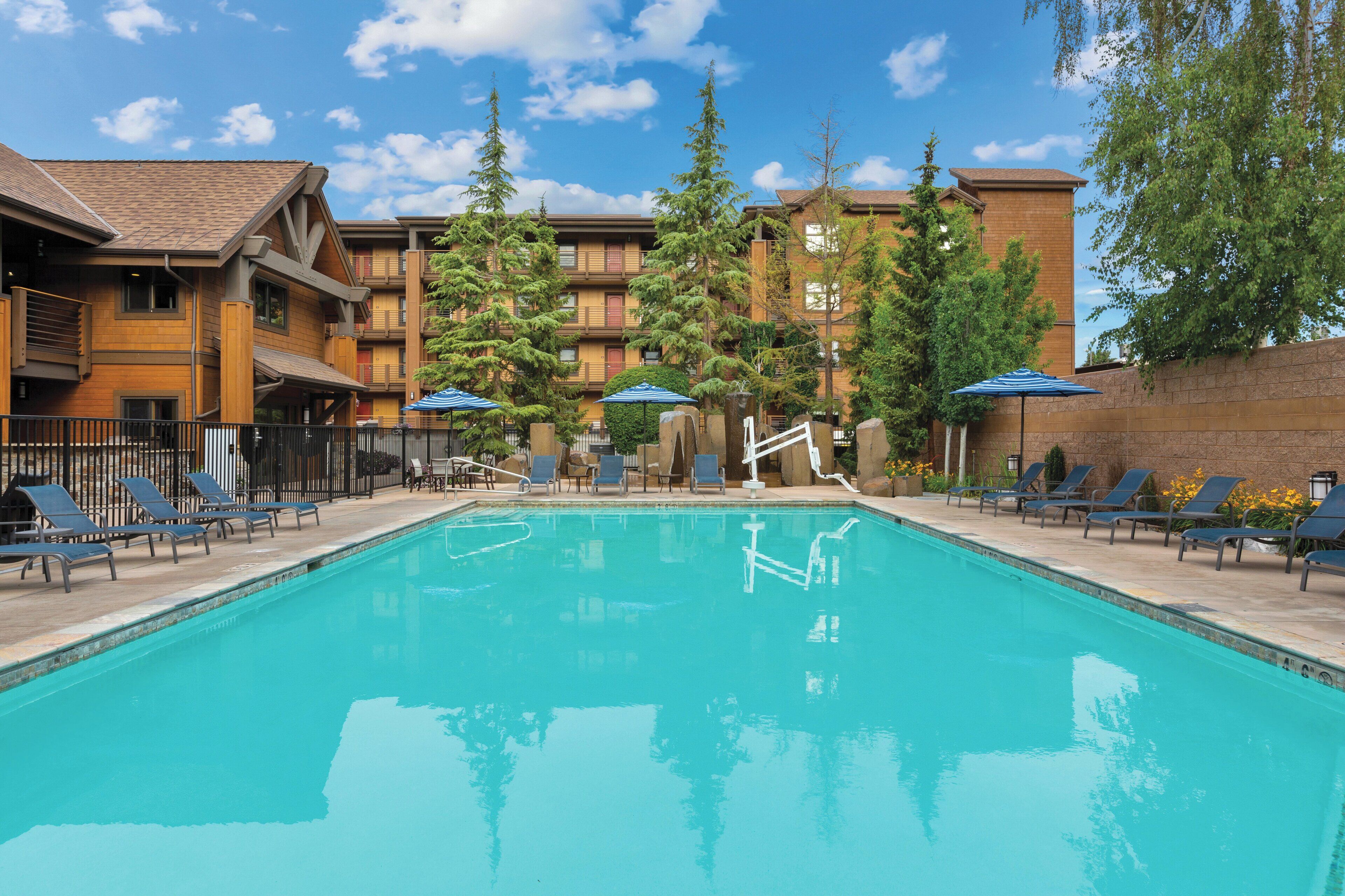 A swimming pool with lounge chairs and umbrellas, surrounded by multi-story buildings with balconies and trees.