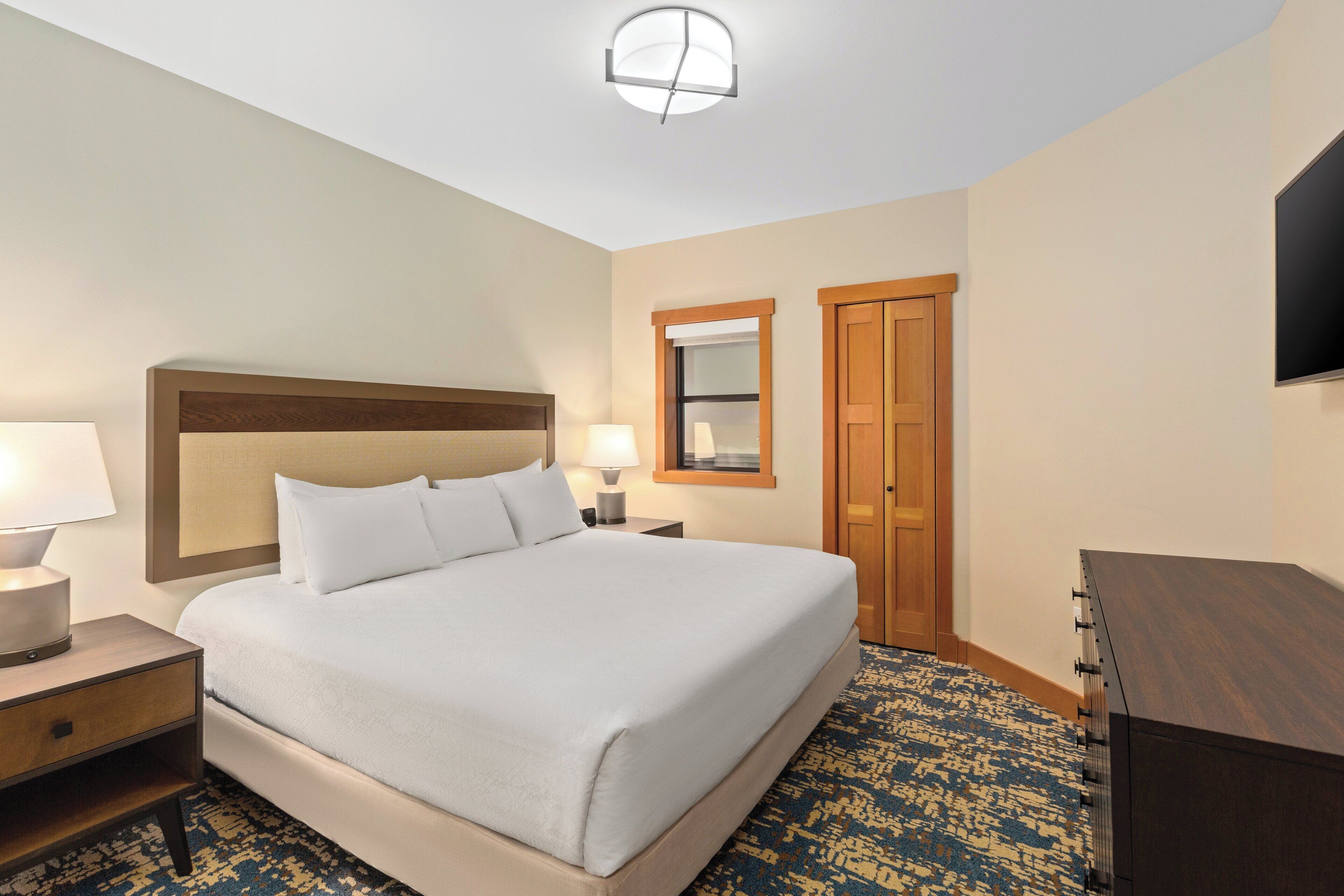 A hotel room with a large bed, bedside lamps, a wooden headboard, a small table, a dresser, a mirror, and a door.