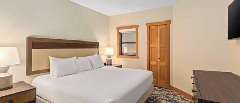 A hotel room with a large bed, bedside lamps, a wooden headboard, a small table, a dresser, a mirror, and a door.