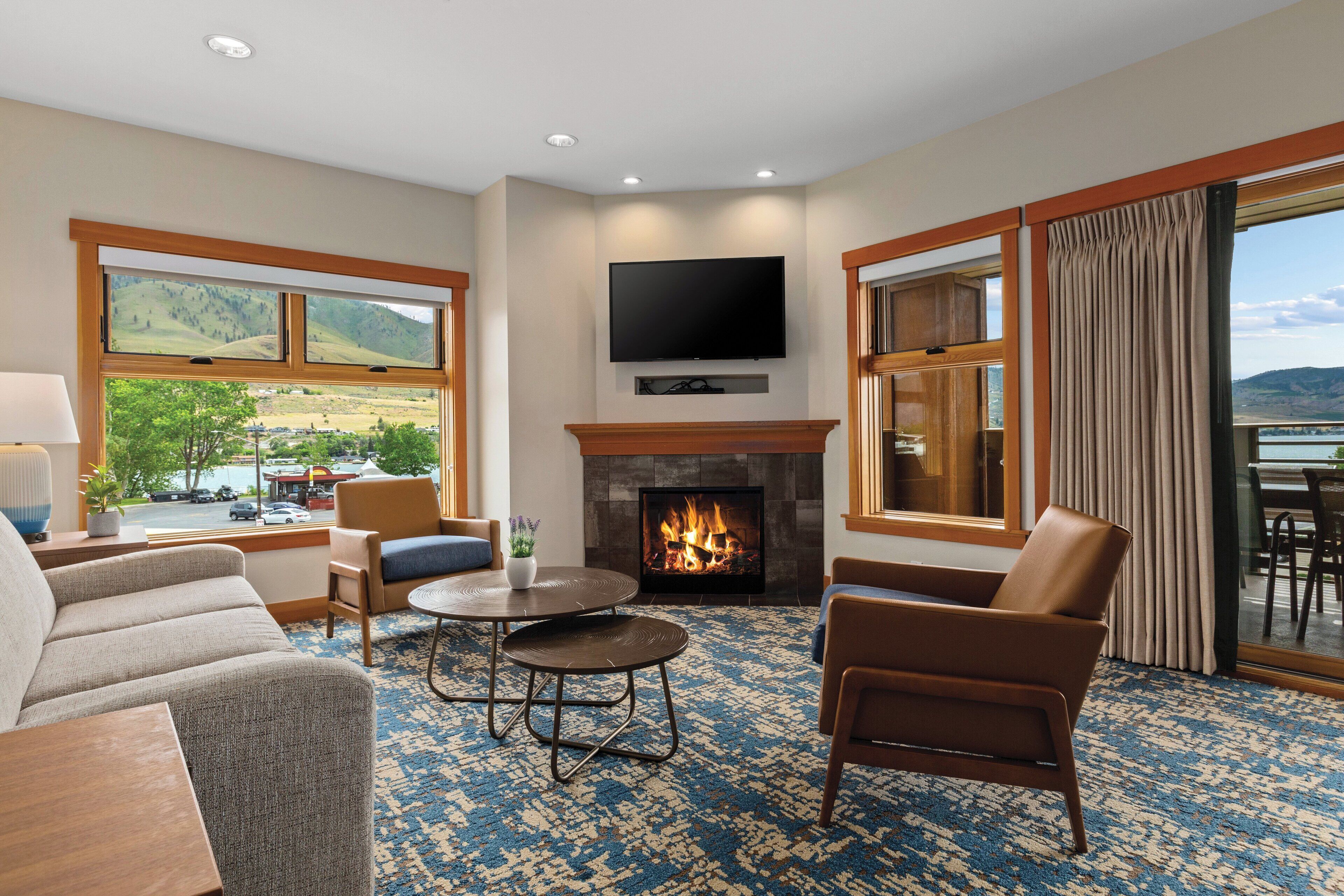 A hotel room with a fireplace, comfortable seating, a flat-screen TV, and large windows offering a view of the outdoors.