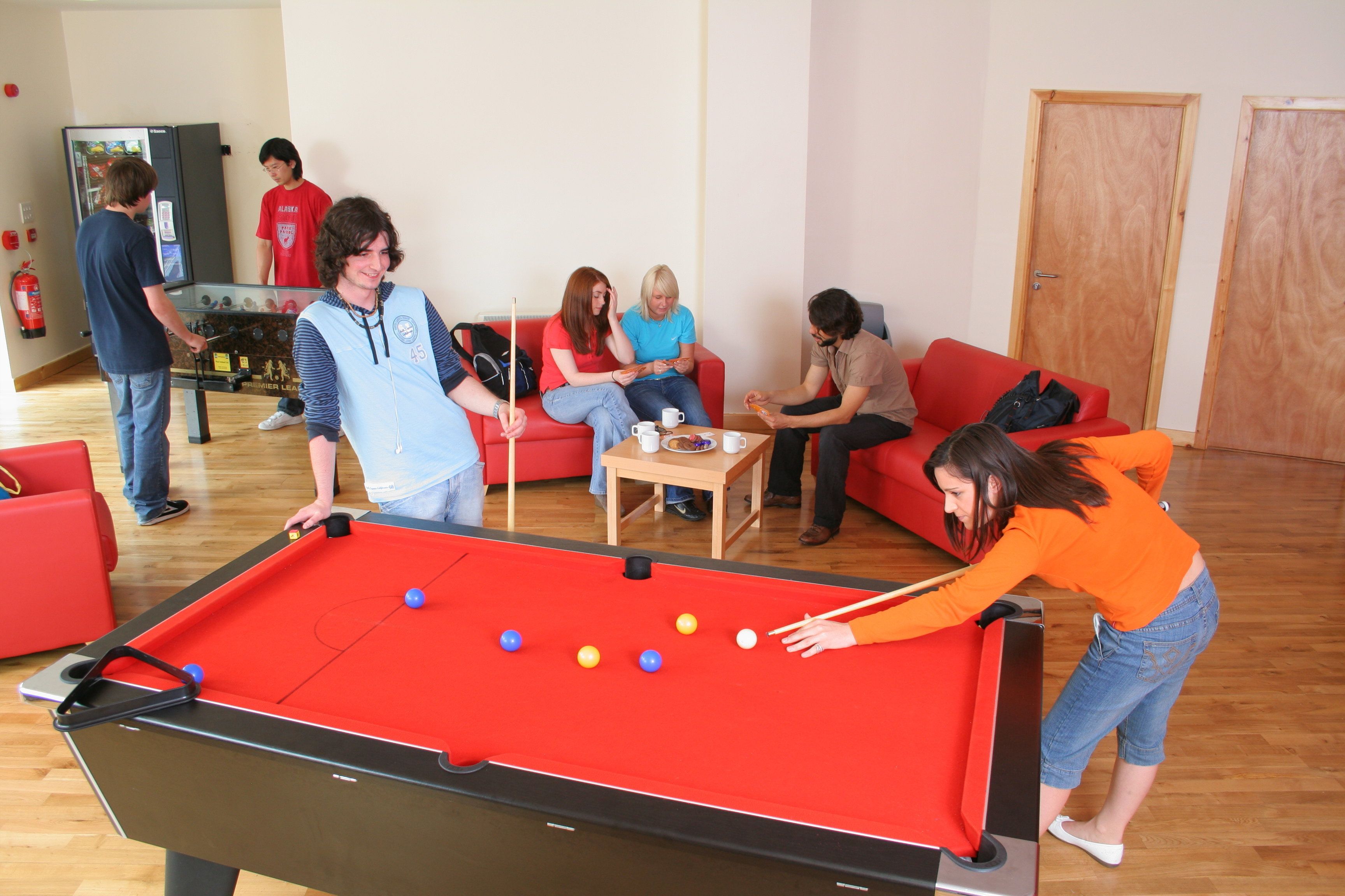 Une table de billard dans une pièce où se trouvent un canapé rouge, une petite table avec des tasses et une porte au fond.