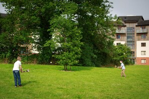 Um campo gramado com duas pessoas brincando, uma árvore grande e um edifício ao fundo.