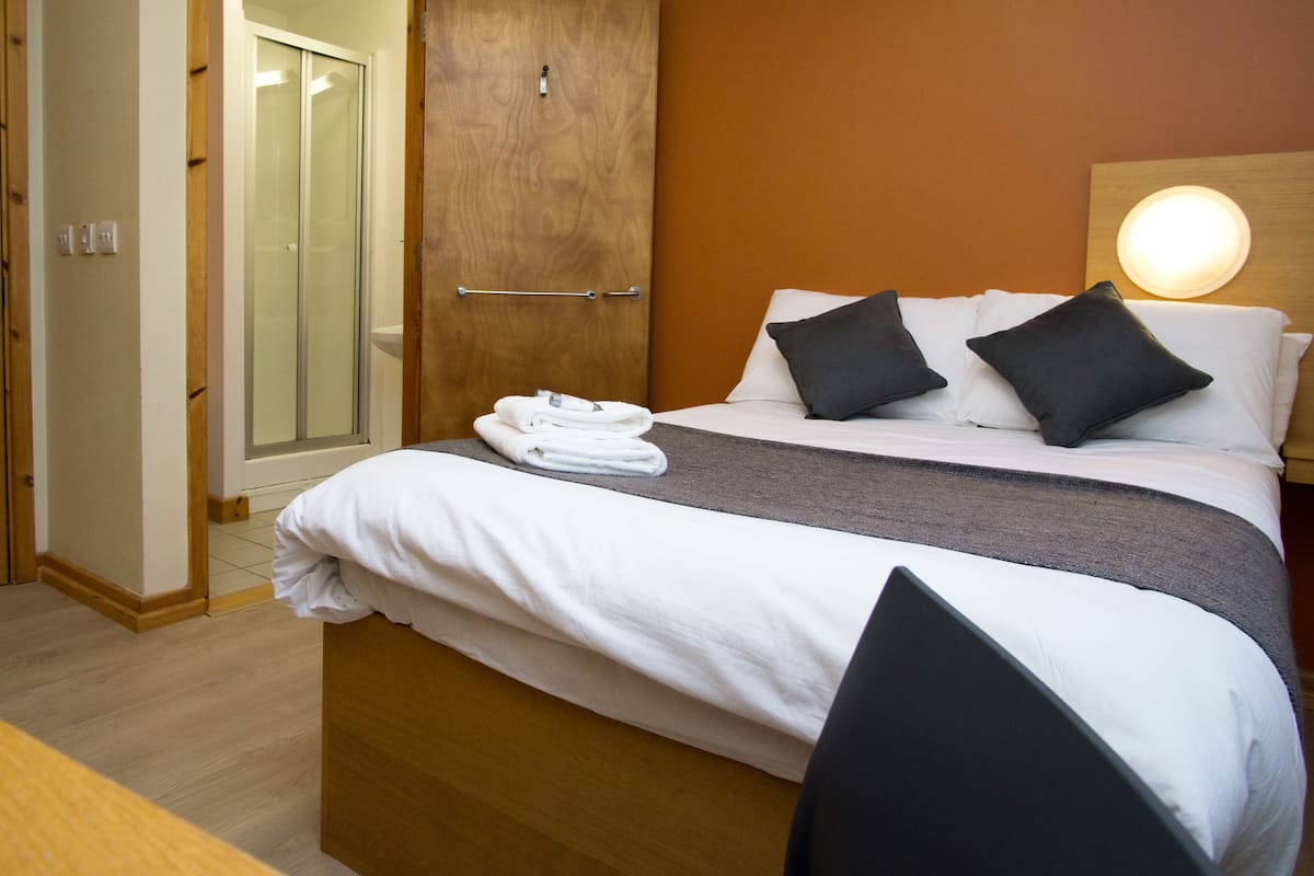 A hotel room with a bed, a wooden wardrobe, a bathroom with a glass door, and a bedside table with towels.