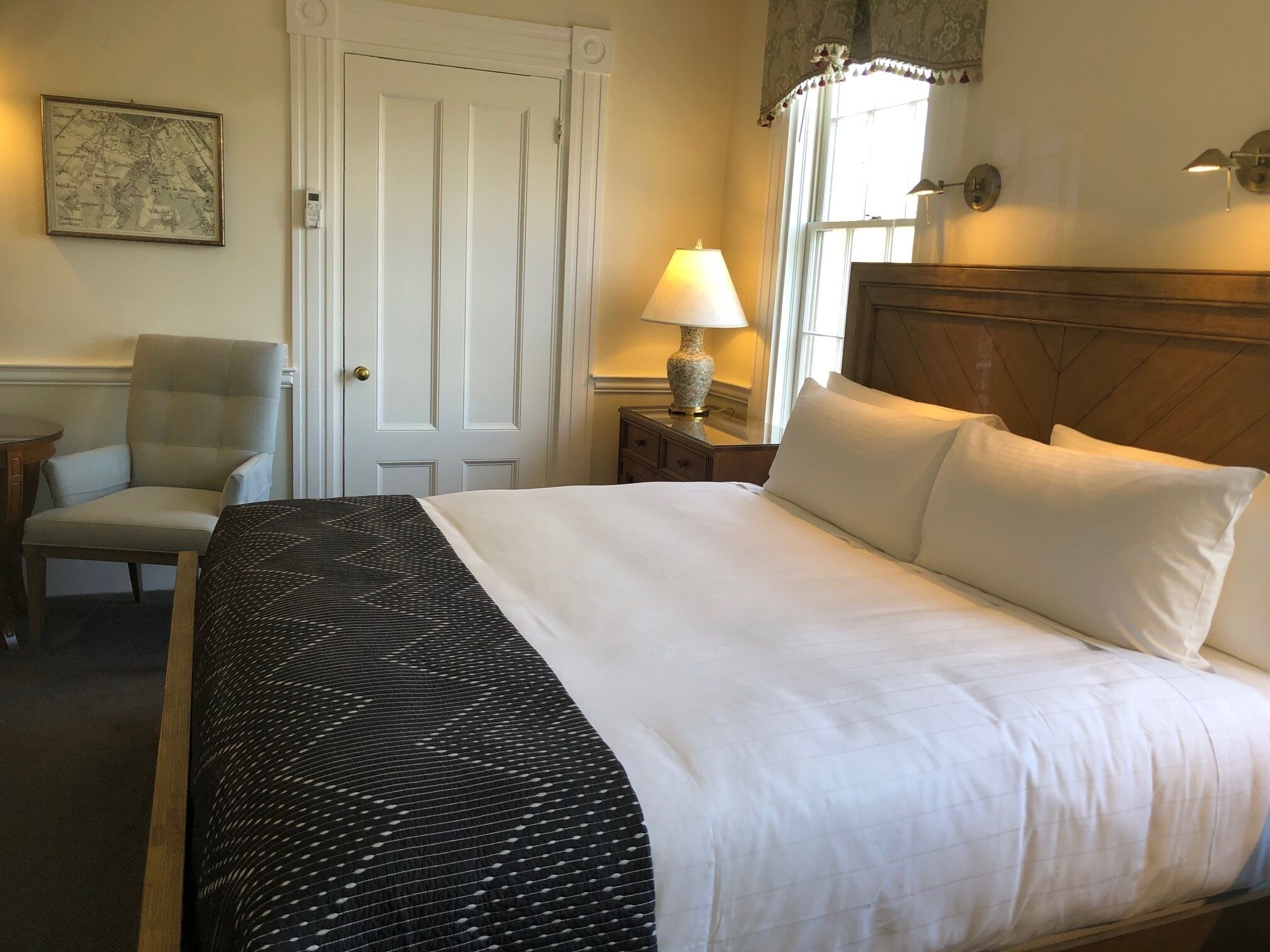 A neatly made bed with white linens, a chair, a lamp, and a map on the wall.