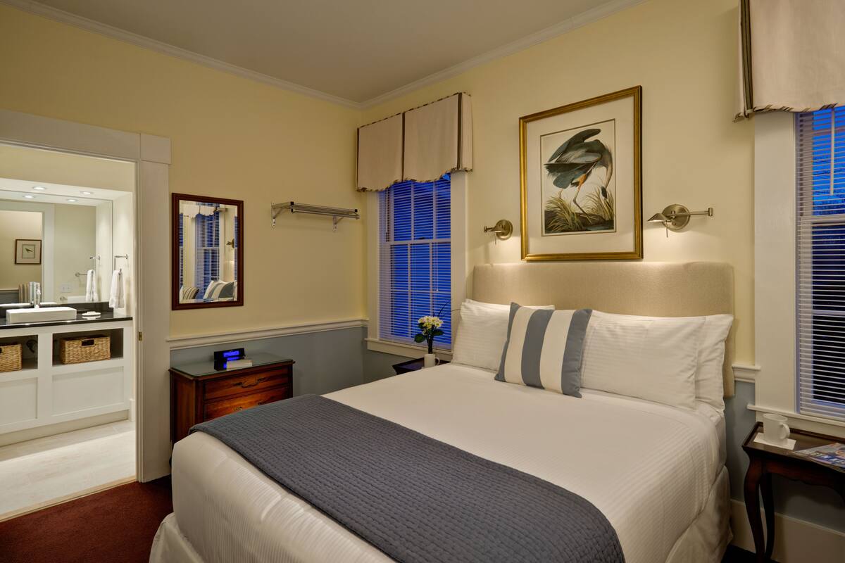 A bedroom with a large bed, a bedside table, a nightstand, a dresser, a mirror, and a window with blinds.