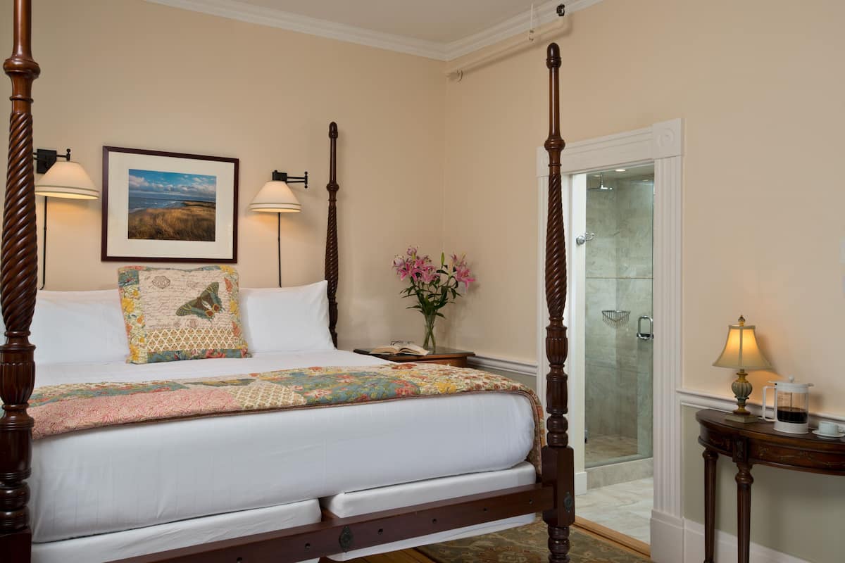 A four-poster bed with a quilt, bedside lamps, a framed picture, a vase with flowers, and a glass-enclosed shower.