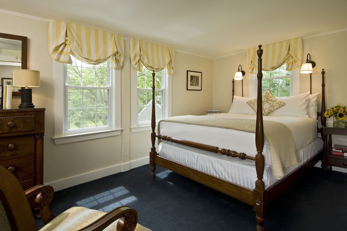 A four-poster bed with a wooden frame, a dark carpeted floor, and a window with yellow-striped valance.