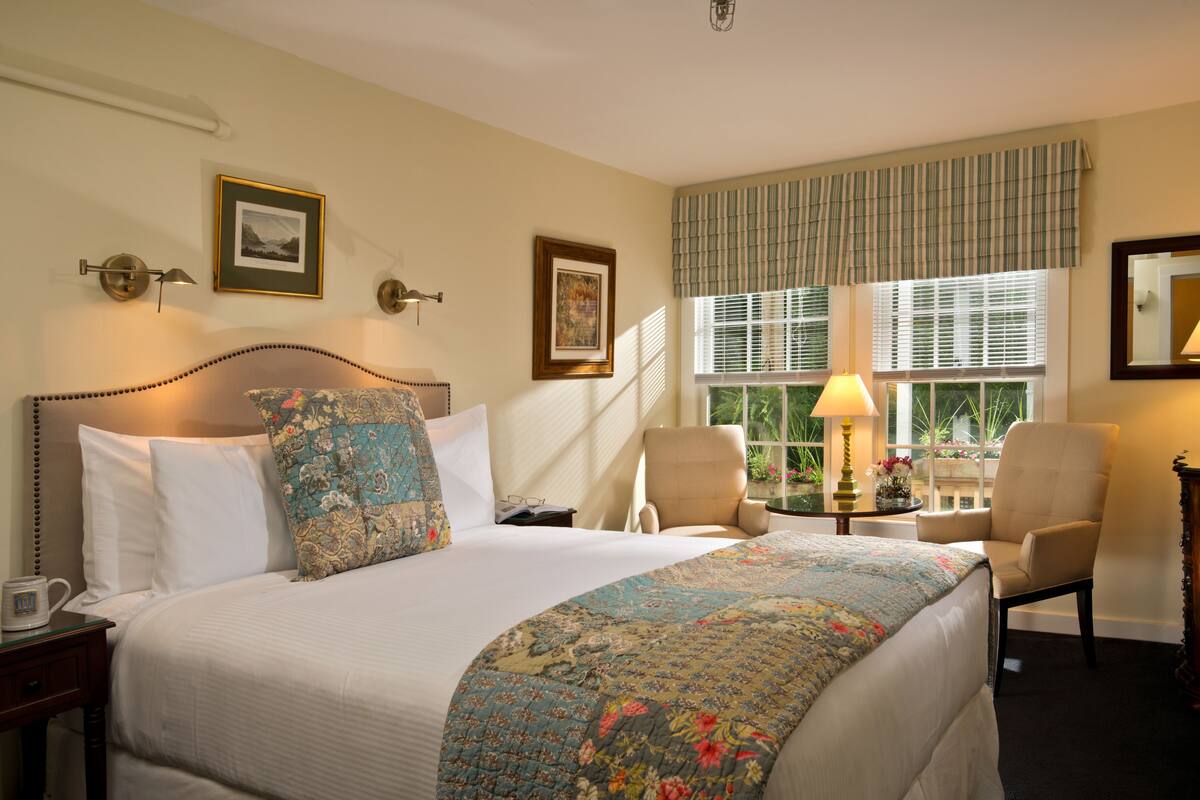 A neatly made bed with a floral quilt, a bedside table with a lamp, and a chair by the window.