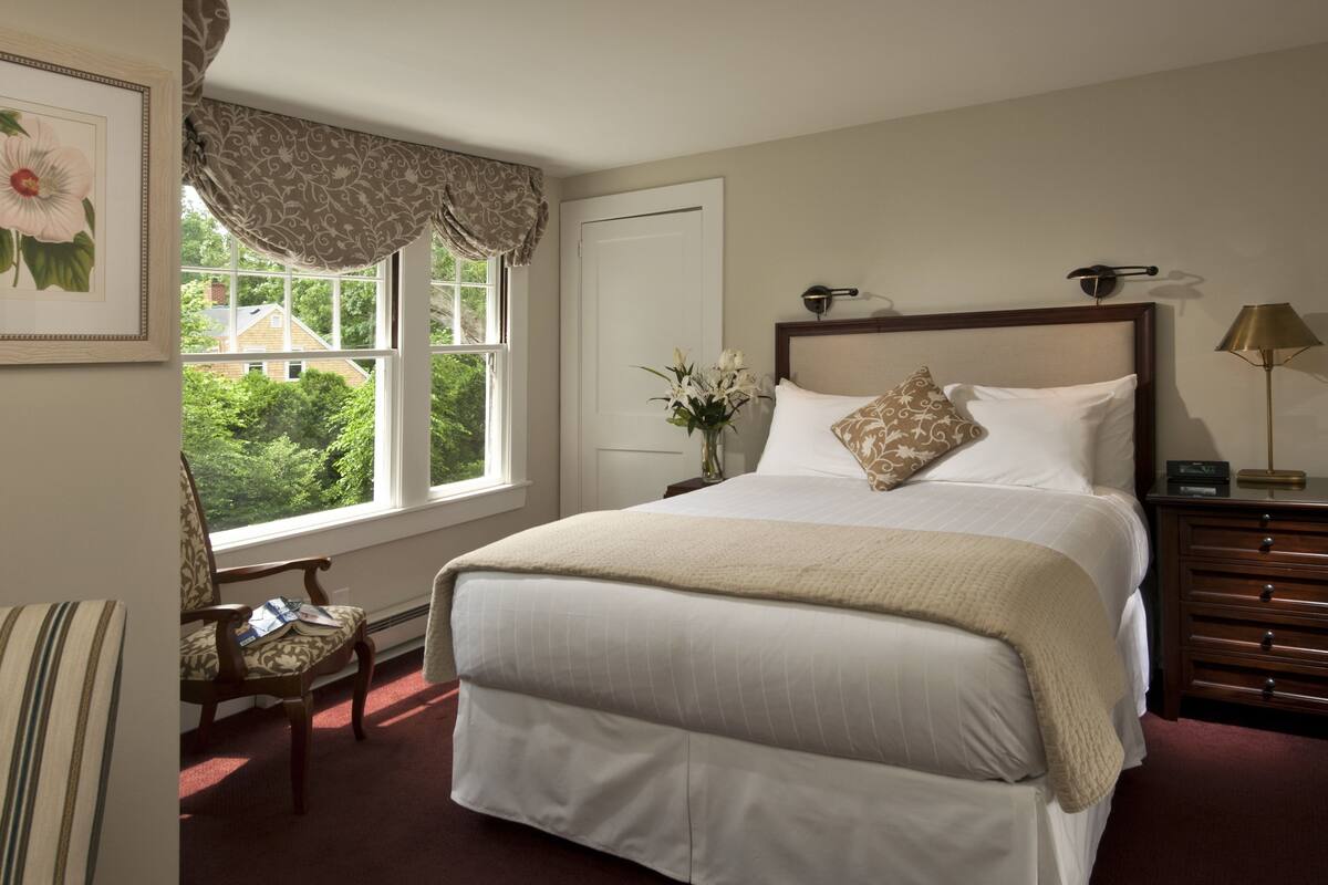 A neatly arranged bedroom with a large bed, a bedside table with a lamp, a chair, and a window with a view of greenery.