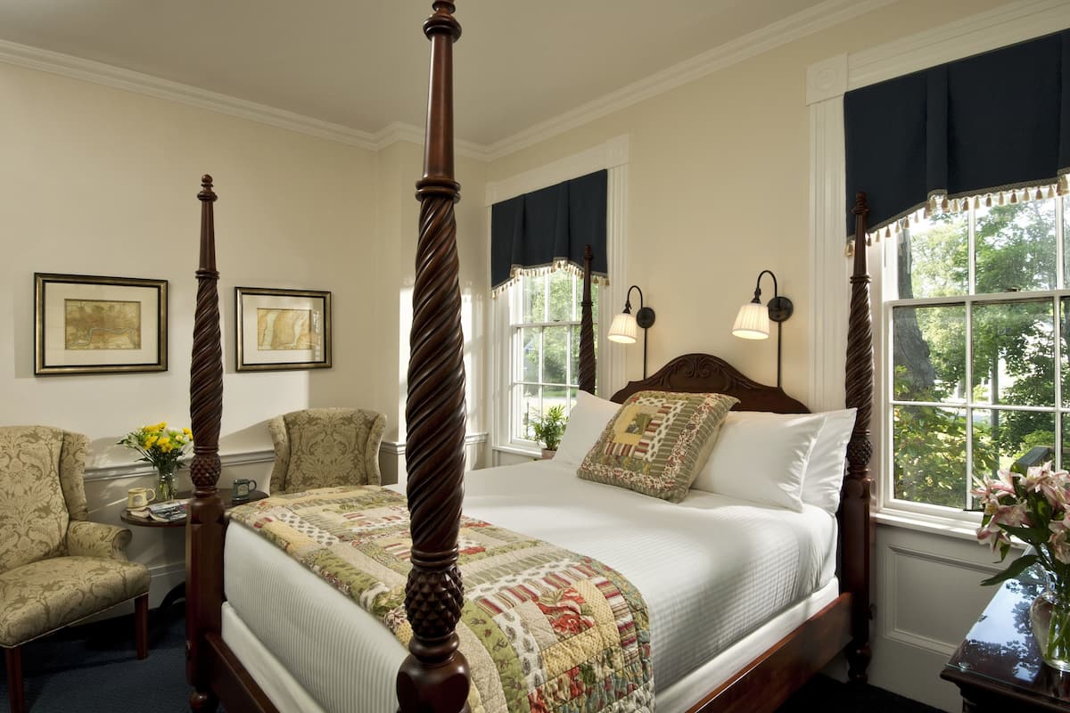 A four-poster bed with a quilt, two armchairs, a small table with a vase of flowers, and a window with a view of trees.