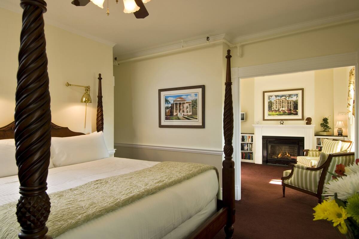 A four-poster bed with a wooden headboard, a fireplace, and a seating area with striped chairs.