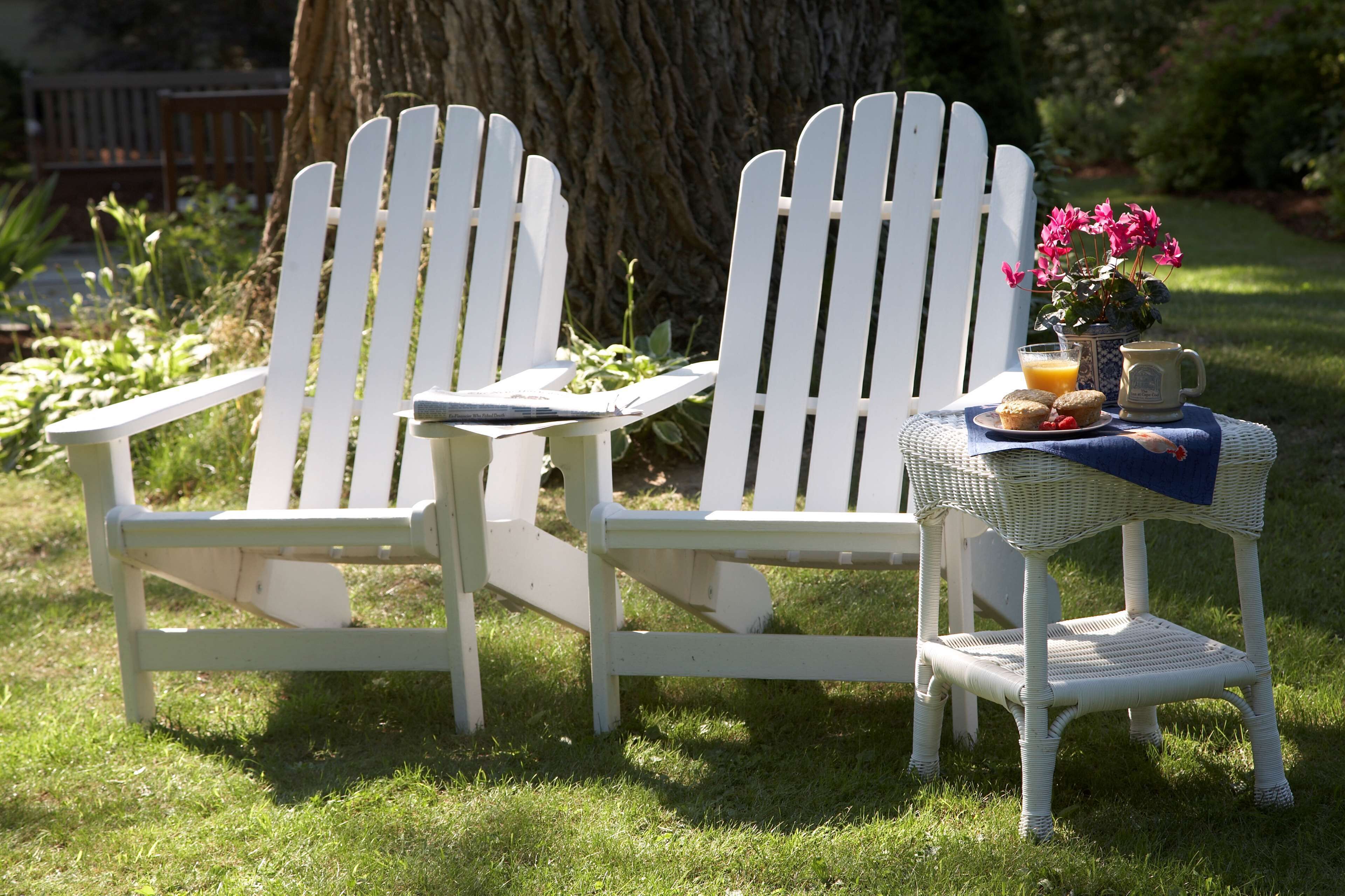 Dos sillas Adirondack blancas con una mesa de mimbre y una planta en maceta entre ellas.