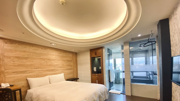 A modern hotel room with a large bed, wooden wall paneling, a small bedside table with a phone, and a view of a cityscape through a glass door. - Burgary Hotel (Taipei)