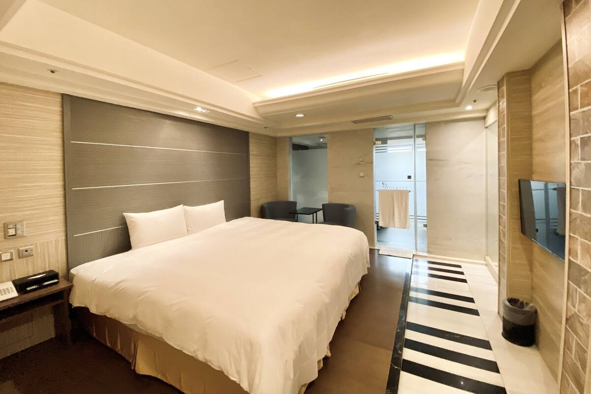 A modern hotel room with a large bed, a flat-screen TV mounted on the wall, a desk with a telephone, and a small seating area with two chairs.