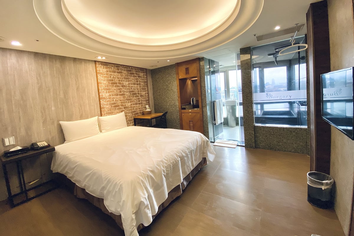 A modern hotel room with a large bed, a wooden headboard, a brick accent wall, a small table with a chair, a vanity with a mirror, and a TV mounted on the wall.