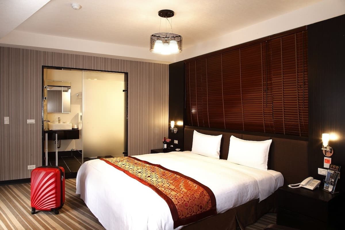 A hotel room with a large bed, a bedside table with a phone, a red suitcase, a mirror, a bathroom with a sink, and a window with blinds.