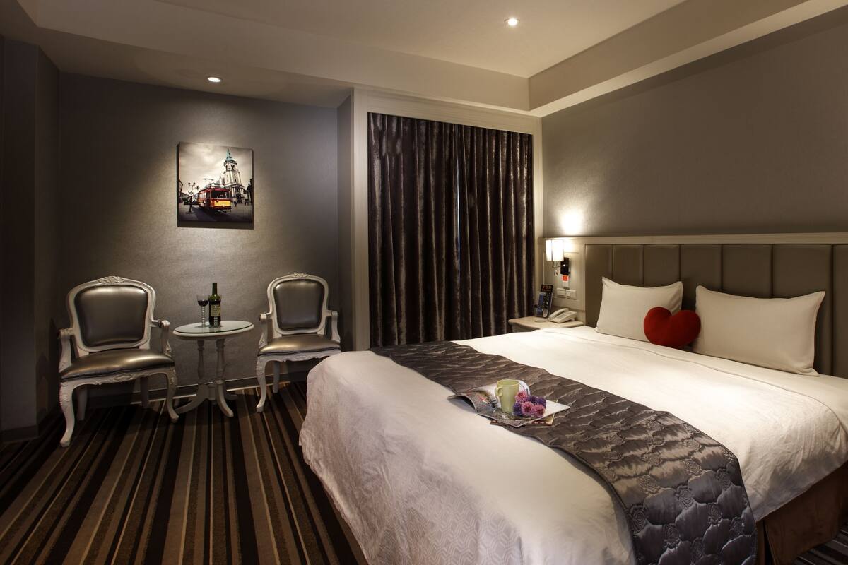 A hotel room with a large bed, two armchairs, a small table, a painting on the wall, and a curtain.