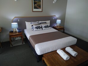 A hotel room with a large bed, two bedside tables with lamps, a wooden bench, and a rolled-up towel on the bench. - Capricorn Motel & Conference Centre (Parkhurst)