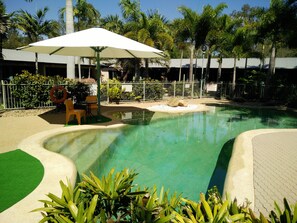 A swimming pool with a white umbrella, surrounded by palm trees and a fenced area with a lifebuoy. - Capricorn Motel & Conference Centre (Parkhurst)