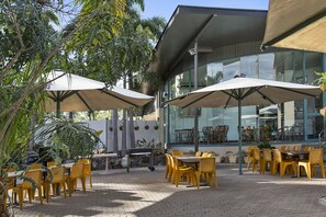 An outdoor patio area with yellow chairs and tables, covered by large umbrellas, and surrounded by potted plants and trees. - Capricorn Motel & Conference Centre (Parkhurst)