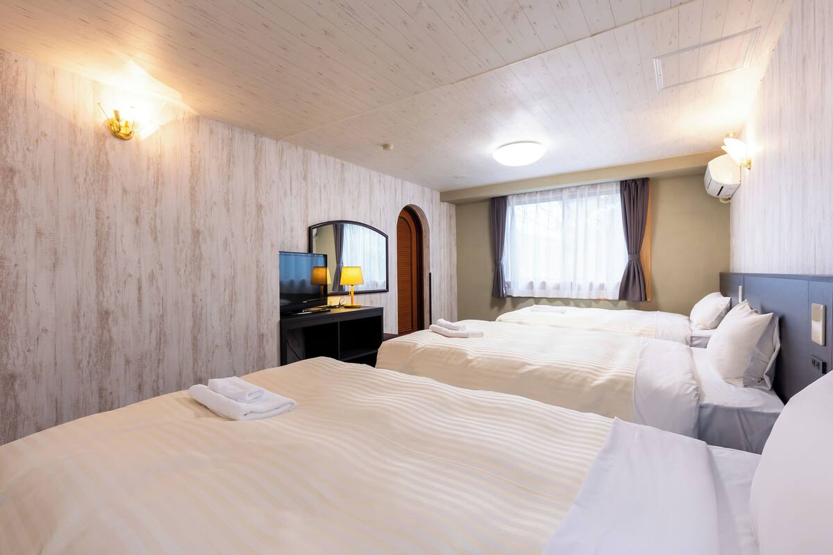 A hotel room with two beds, a desk with a lamp, a mirror, and a window with curtains.