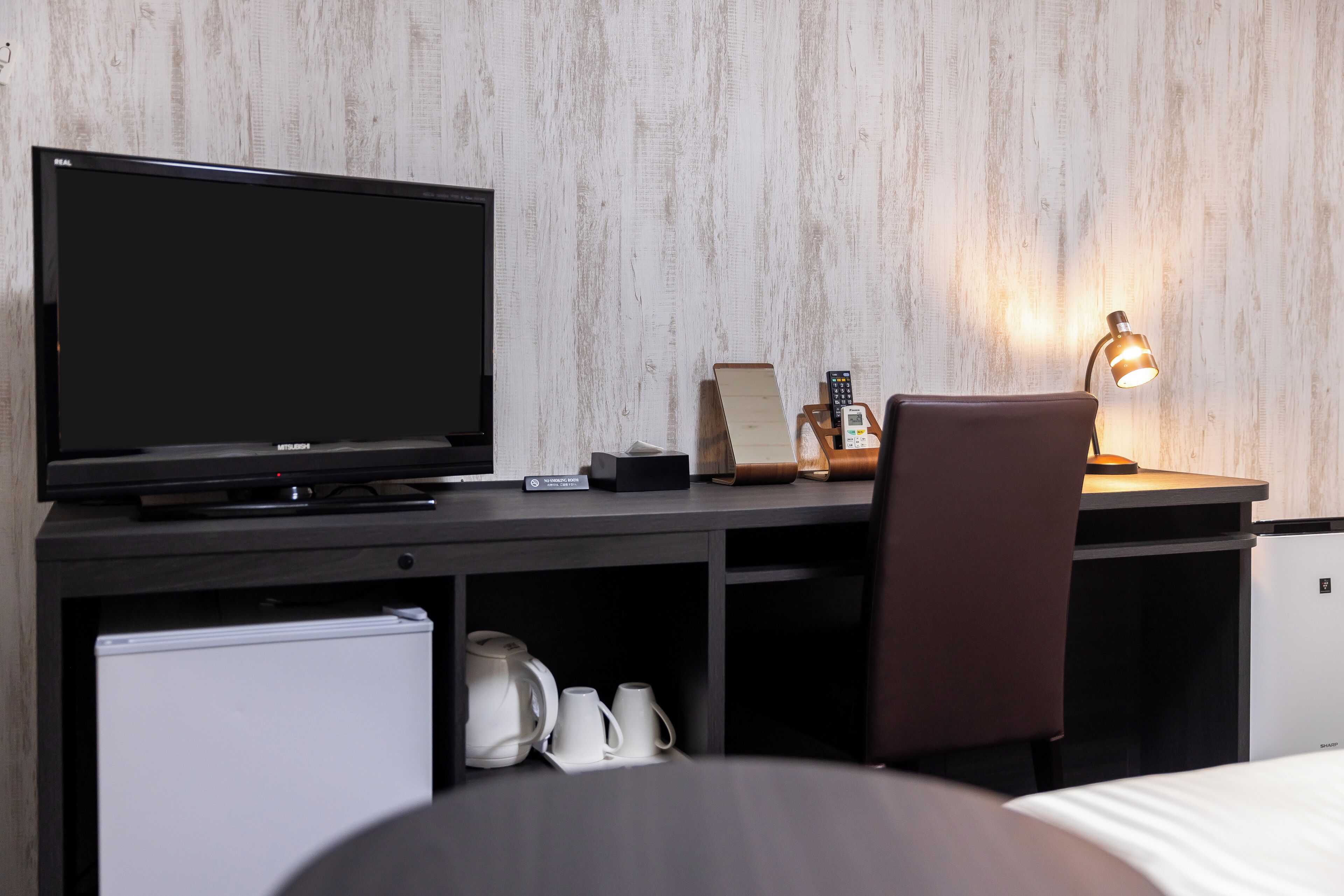 A hotel room with a flat-screen TV, a desk with a lamp, a chair, a mini-fridge, and a coffee maker.