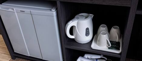 Una cocina compacta con un refrigerador Haier, un hervidor eléctrico blanco, dos tazas blancas y un cargador portátil.