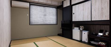 Ein Zimmer mit Tatami-Boden, einer verschiebbaren Shoji-Schirmwand, einer Küchenzeile mit Kühlschrank und Mikrowelle sowie einem kleinen Regal mit Gegenständen.