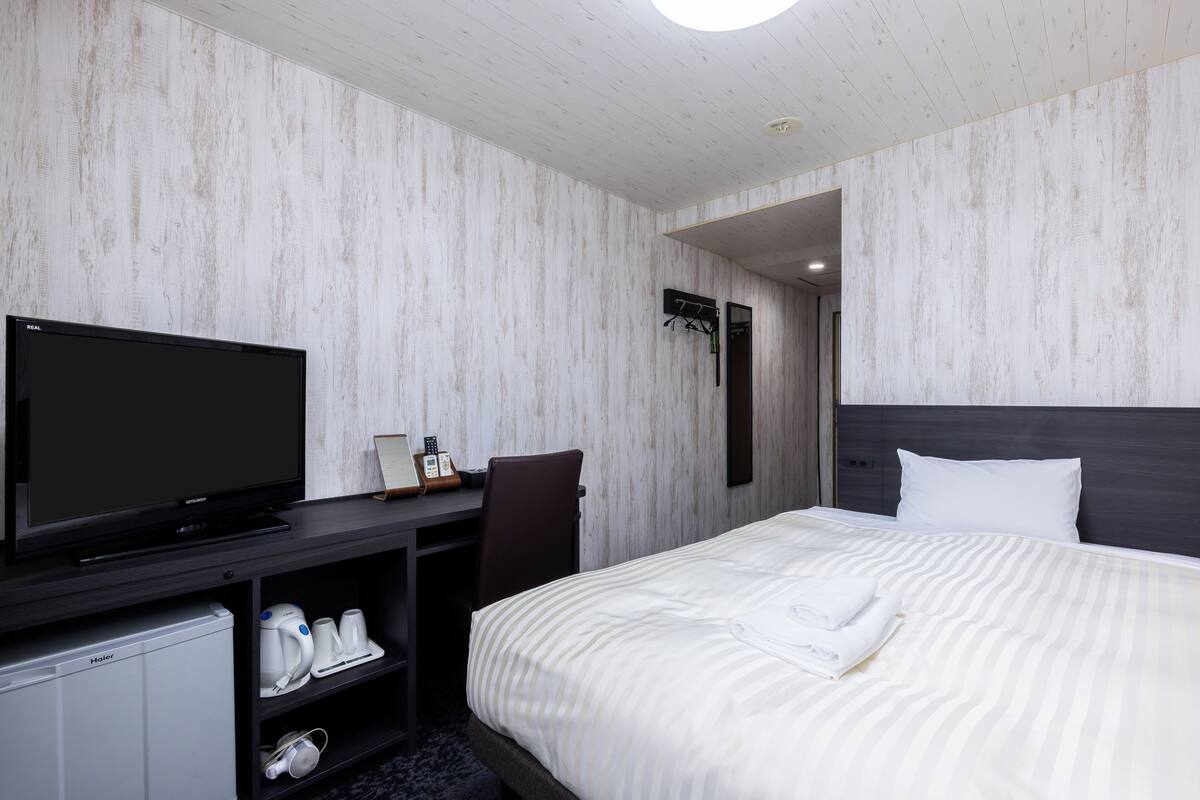 A hotel room with a bed, a television, a desk with a chair, a small refrigerator, and a coat rack.