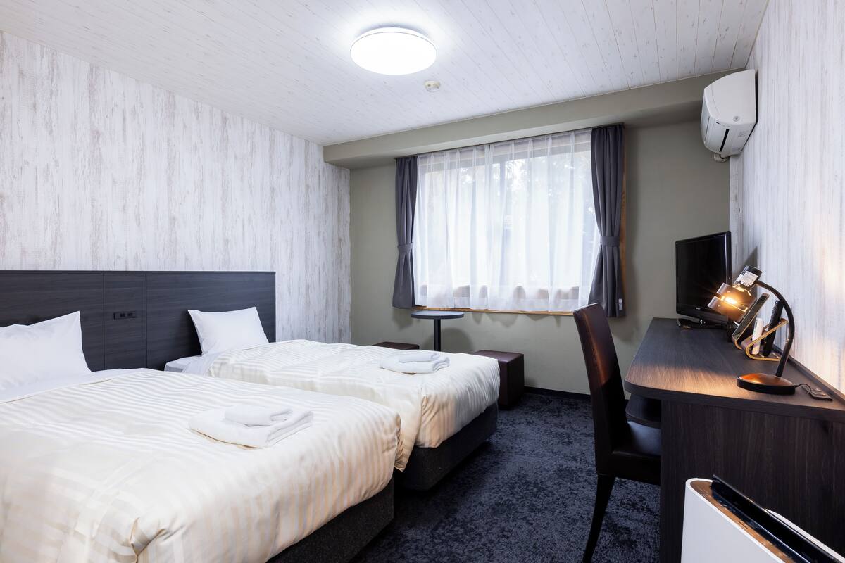 A hotel room with two beds, a desk with a lamp, a chair, a TV, and an air conditioning unit.