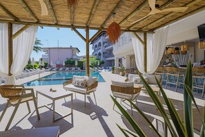 A poolside area with lounge chairs, a bar, and a view of a building in the background. - Hotel Tropical (Kassandra)