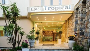 La entrada del Hotel Tropical, con fachada de piedra, plantas en macetas y un interior bien iluminado visible a través de puertas de vidrio.