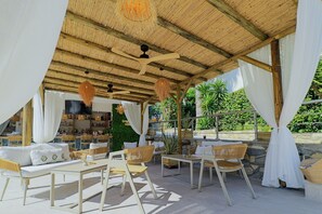 An outdoor seating area with wicker furniture, a thatched roof, and a view of greenery. - Hotel Tropical (Kassandra)