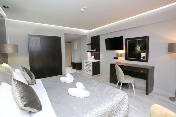 A modern hotel room with a large bed, a desk, a chair, a wardrobe, a flat-screen TV, and a small kitchenette.
