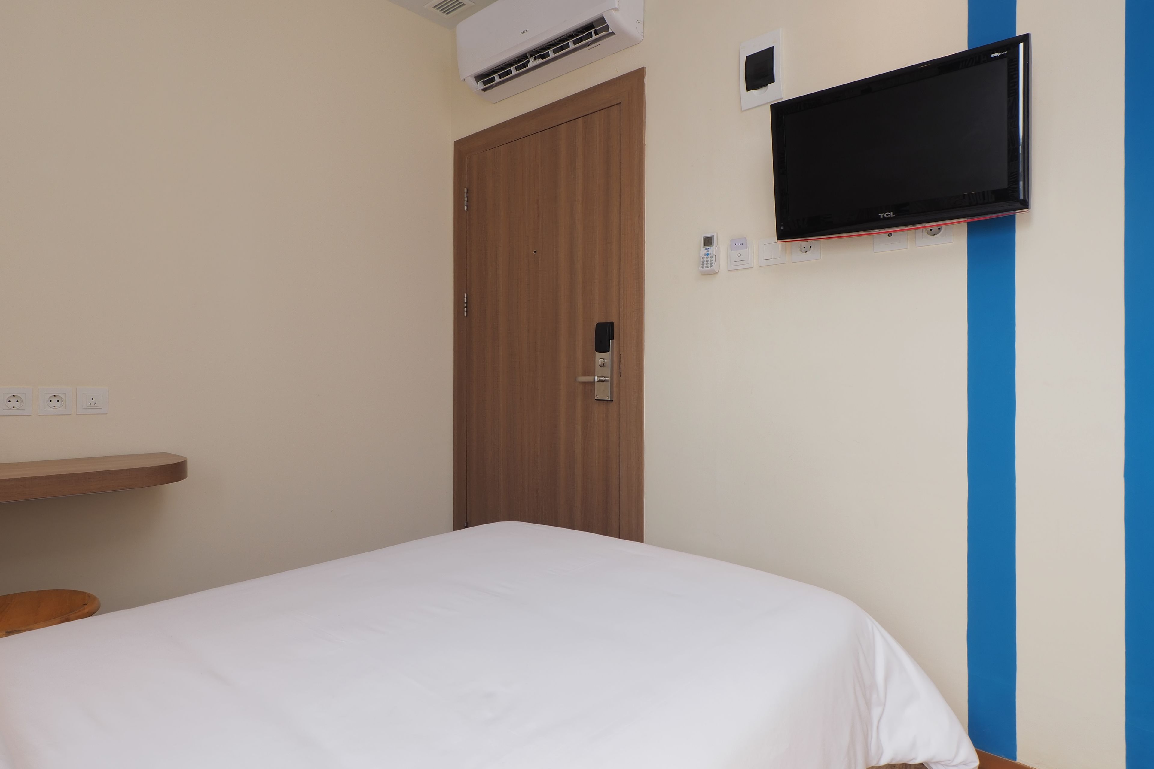 Un dormitorio con una cama, un televisor montado en la pared, una puerta y un enchufe.