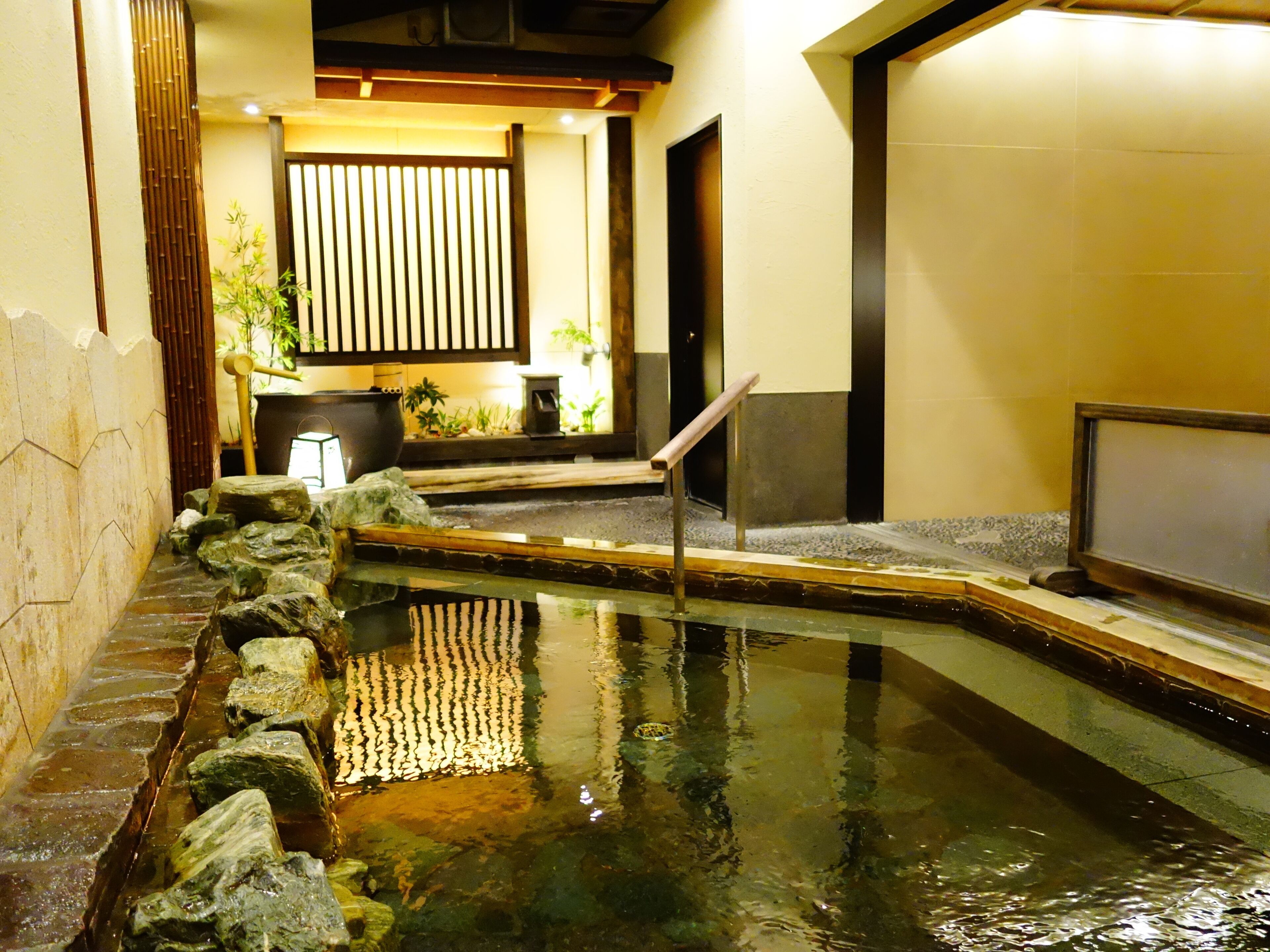 Un onsen japonés tradicional con un sendero de piedra que conduce a una pequeña piscina, rodeada de rocas y una cerca de madera.