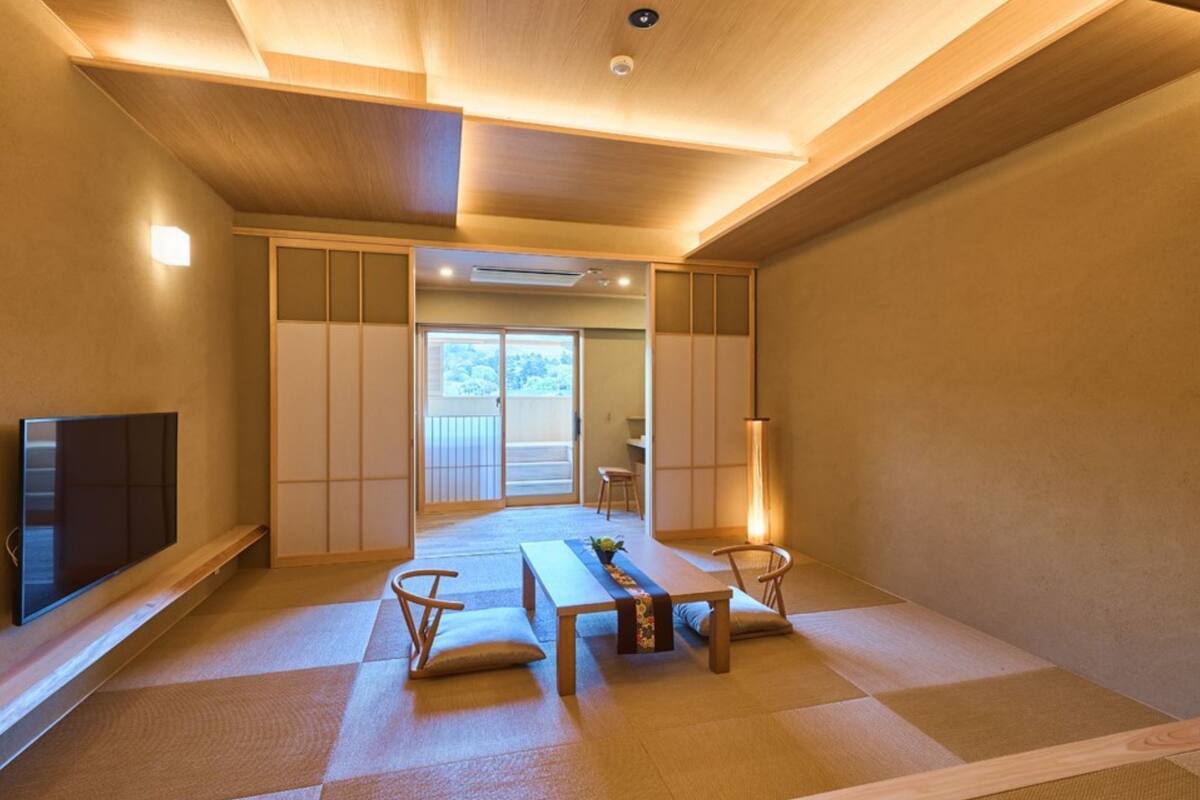 A traditional Japanese-style room with tatami flooring, a low table with cushions, a television, sliding doors, and a view of a balcony.