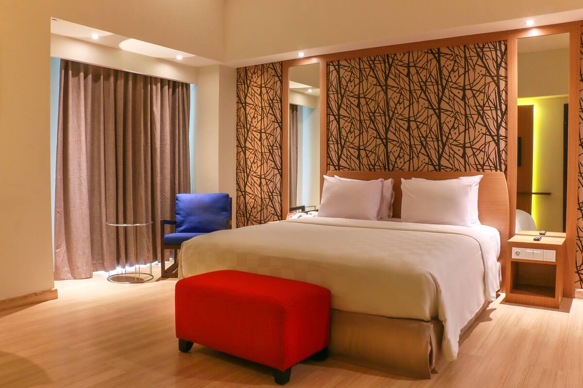 A hotel room with a large bed, a red bench, a blue chair, and a wooden nightstand.