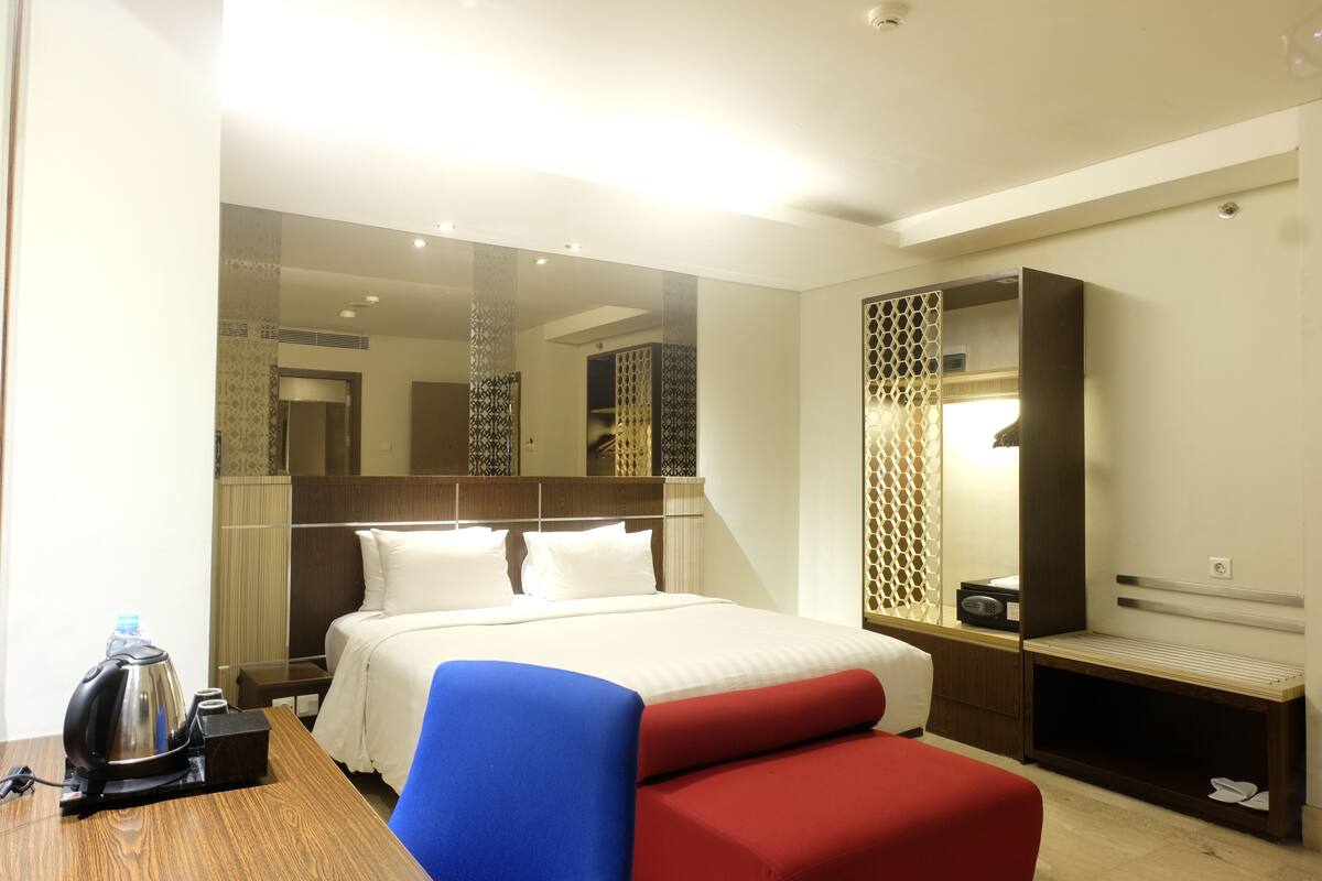 A hotel room with a bed, a desk with a kettle, a chair, a bench, and a mirror.