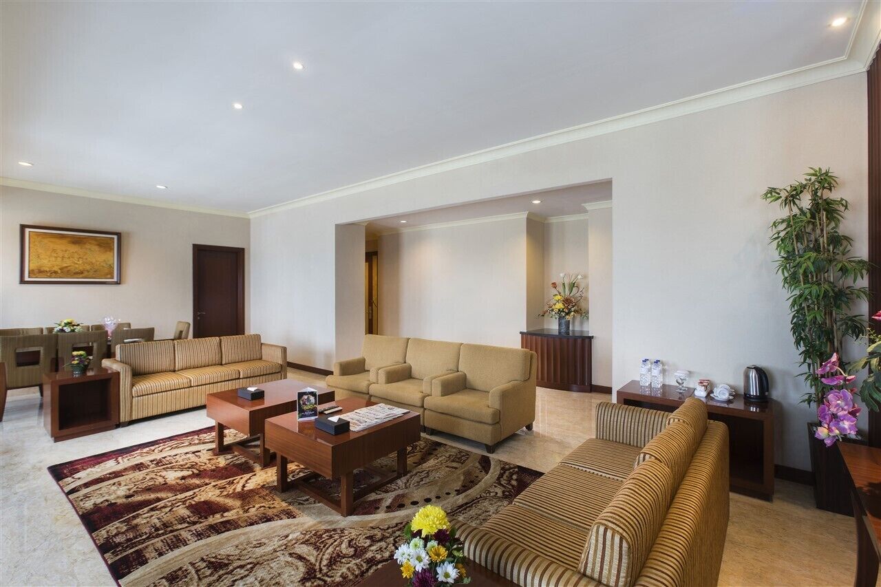 Amplia sala de estar con sofá, sillones, una mesa de centro y una alfombra. Hay una planta y una mesita con objetos decorativos.