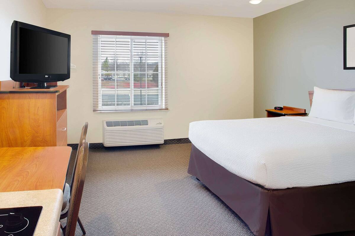 A hotel room with a bed, a desk with a computer, a television, a window with blinds, and an air conditioning unit.