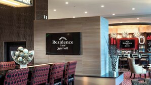 A hotel lobby with a reception desk, bar area, seating with patterned chairs, and a large TV displaying the hotel name. - Residence Inn Boston Needham (Needham)