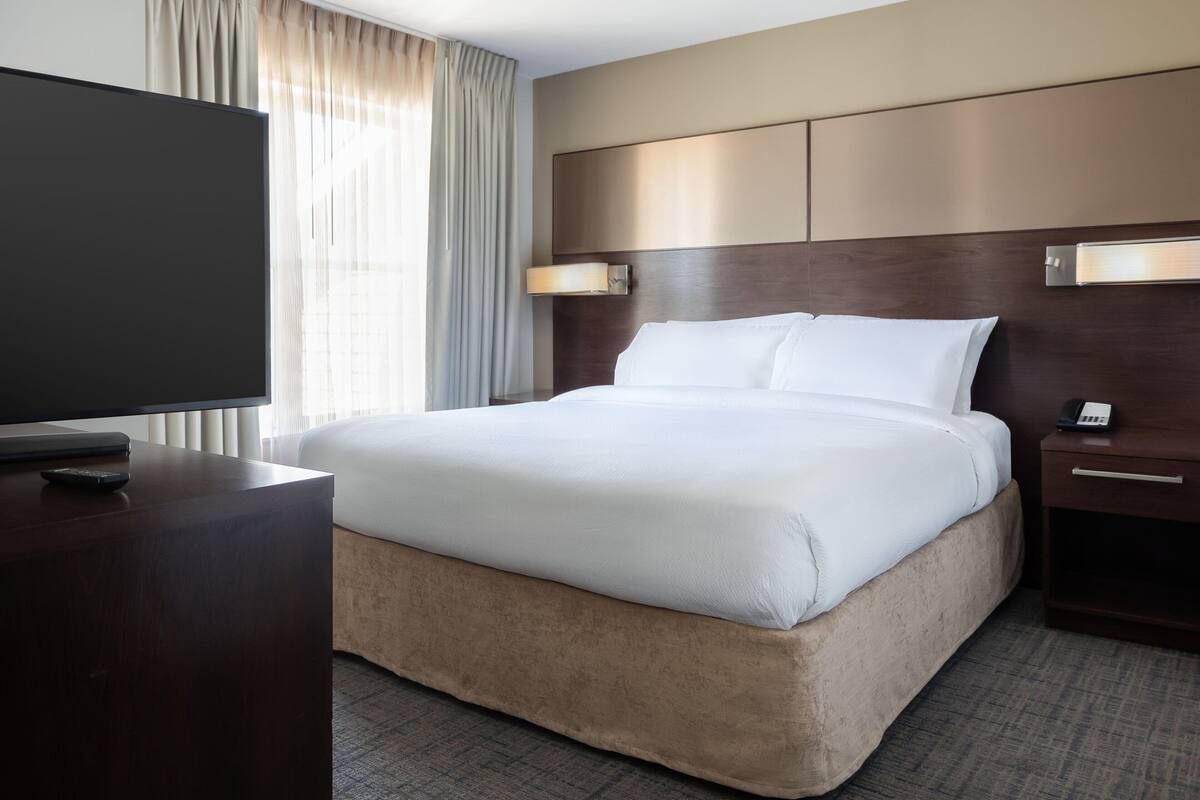 A hotel room with a large bed, a flat-screen TV on a wooden side table, and a bedside table with a lamp.