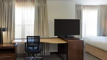 Ein Hotelzimmer mit einem Holzbüro, einem schwarzen Bürostuhl, einem Flachbildfernseher, einem Bett und beigen Vorhängen.
