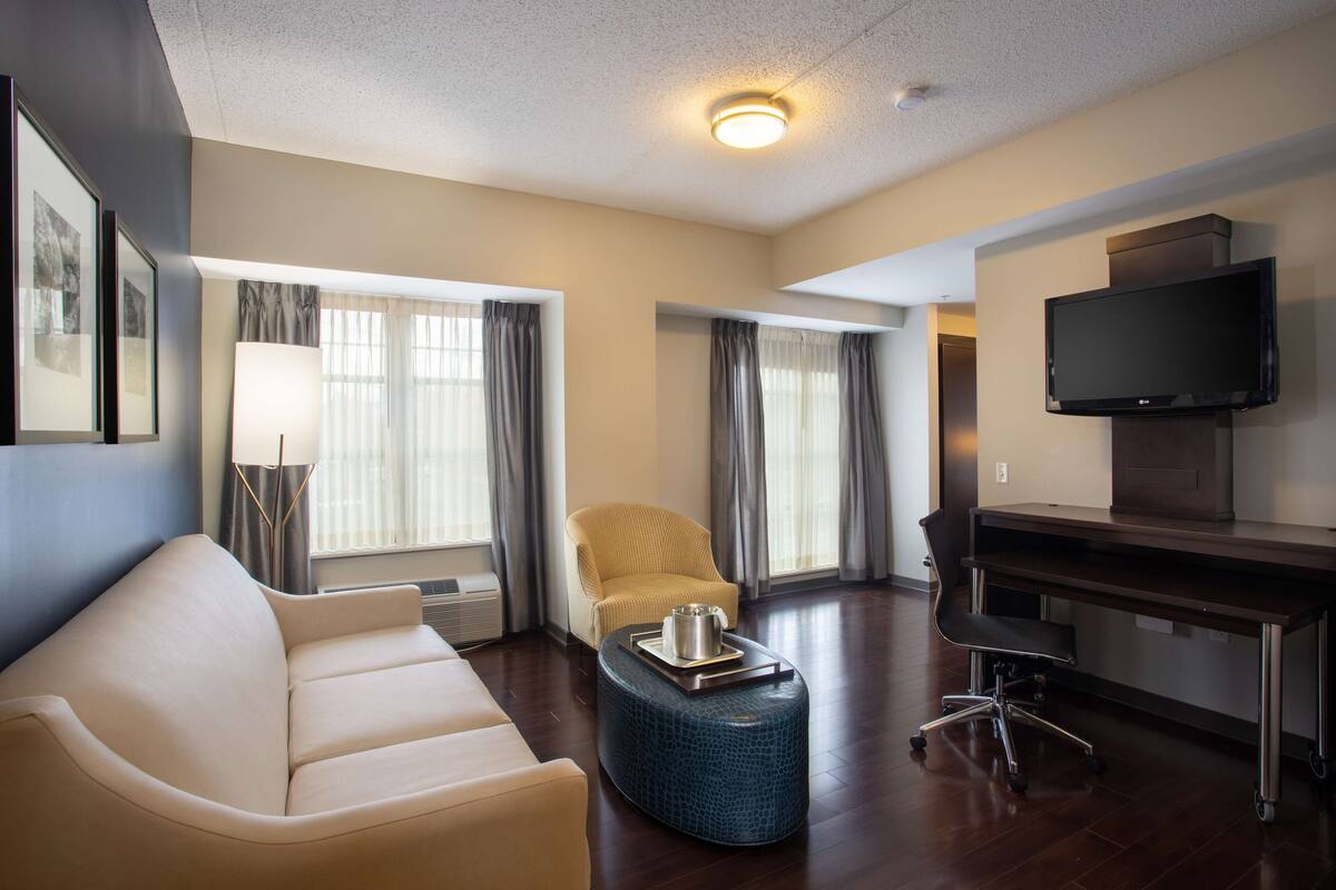 Una habitación de hotel moderna con sofá, sillón, reposapiés, escritorio y televisor.