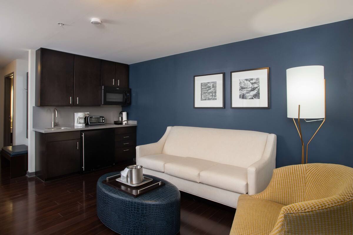 Una sala de estar moderna con un sofá blanco, un reposapiés azul, un sillón amarillo, una zona de cocina con armarios oscuros y un microondas, y dos cuadros enmarcados en la pared.